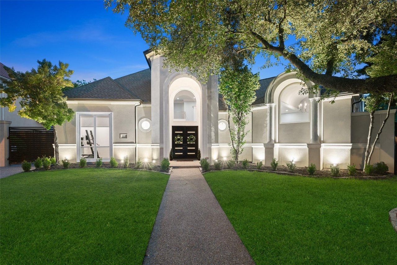 Gray stucco home with arched entry, broad lawn, and mature trees framing the front walk.