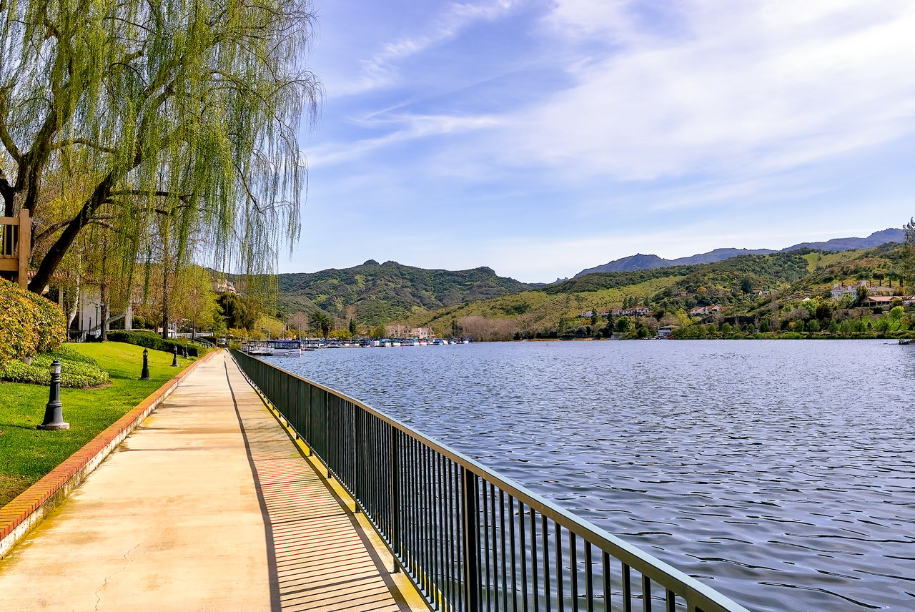 Lakeside walkway beside a broad lake, with willow trees and green hills dotted with homes.