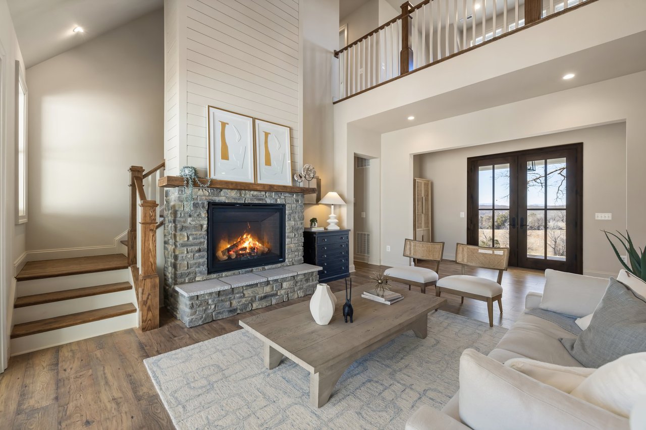 Living room with a stone fireplace, vaulted ceiling, wood staircase, and French doors overlooking trees.