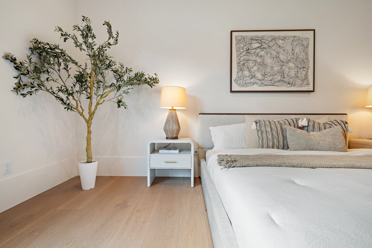 Furnished bedroom with a white bed, potted tree, light wood floors, and framed abstract art.