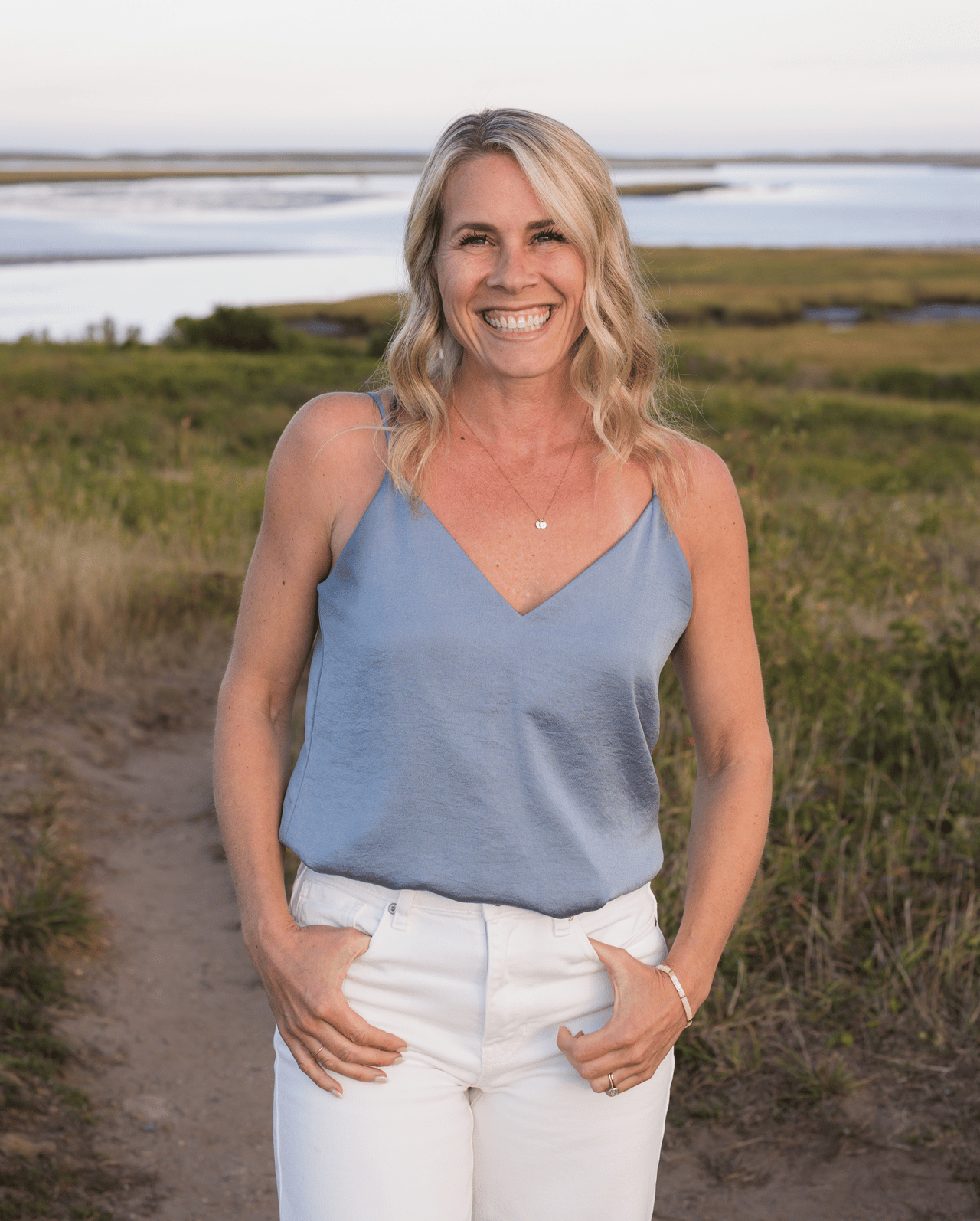 Smiling blonde woman in a blue top and white pants beside a coastal marsh.