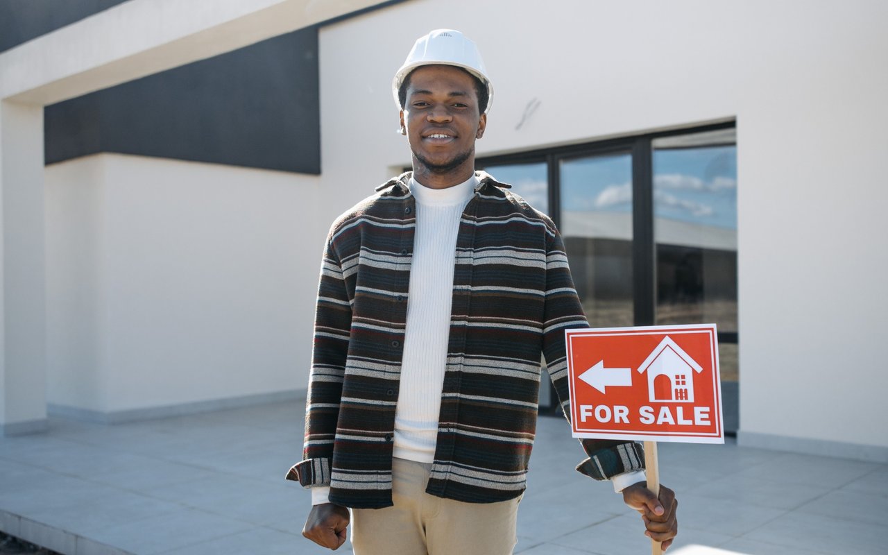 Man in a white hard hat holding a red FOR SALE sign outside a modern house.