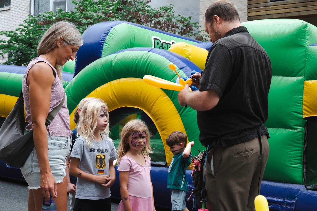 Children with face paint watch a man make a balloon animal beside a colorful bounce house.