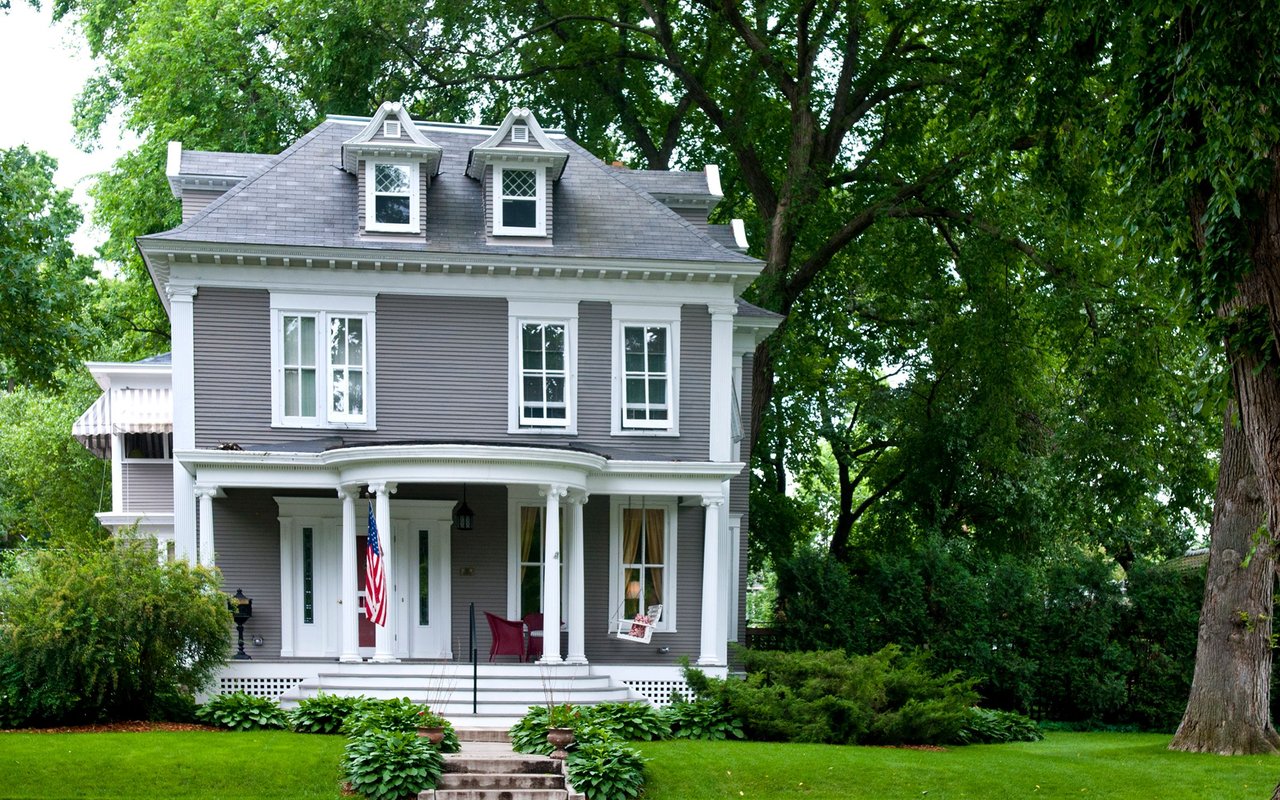 Gray two-story house with white trim, a columned porch, and leafy trees surrounding the lawn.