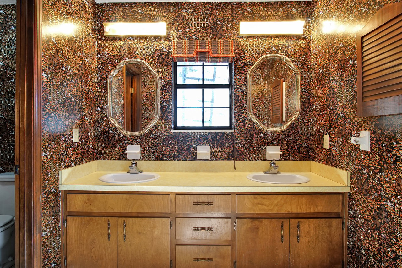 Vintage bathroom with bold, intricate wallpaper in brown tones, dual sinks on a yellow countertop, a centered window, and oval mirrors, creating a retro vibe.