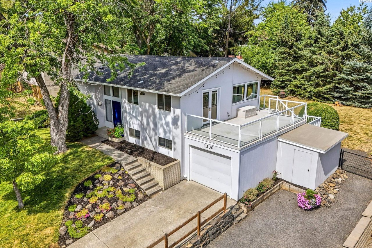 White house with a raised deck and attached garage, surrounded by mature trees and landscaped gardens.