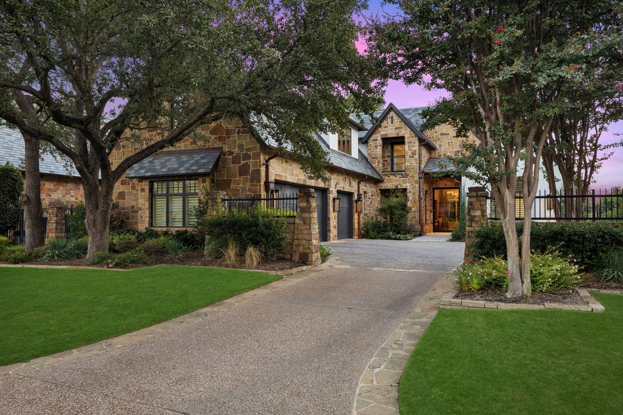 Stone house with a lit entrance, mature trees, landscaped lawn, and curved driveway at dusk.