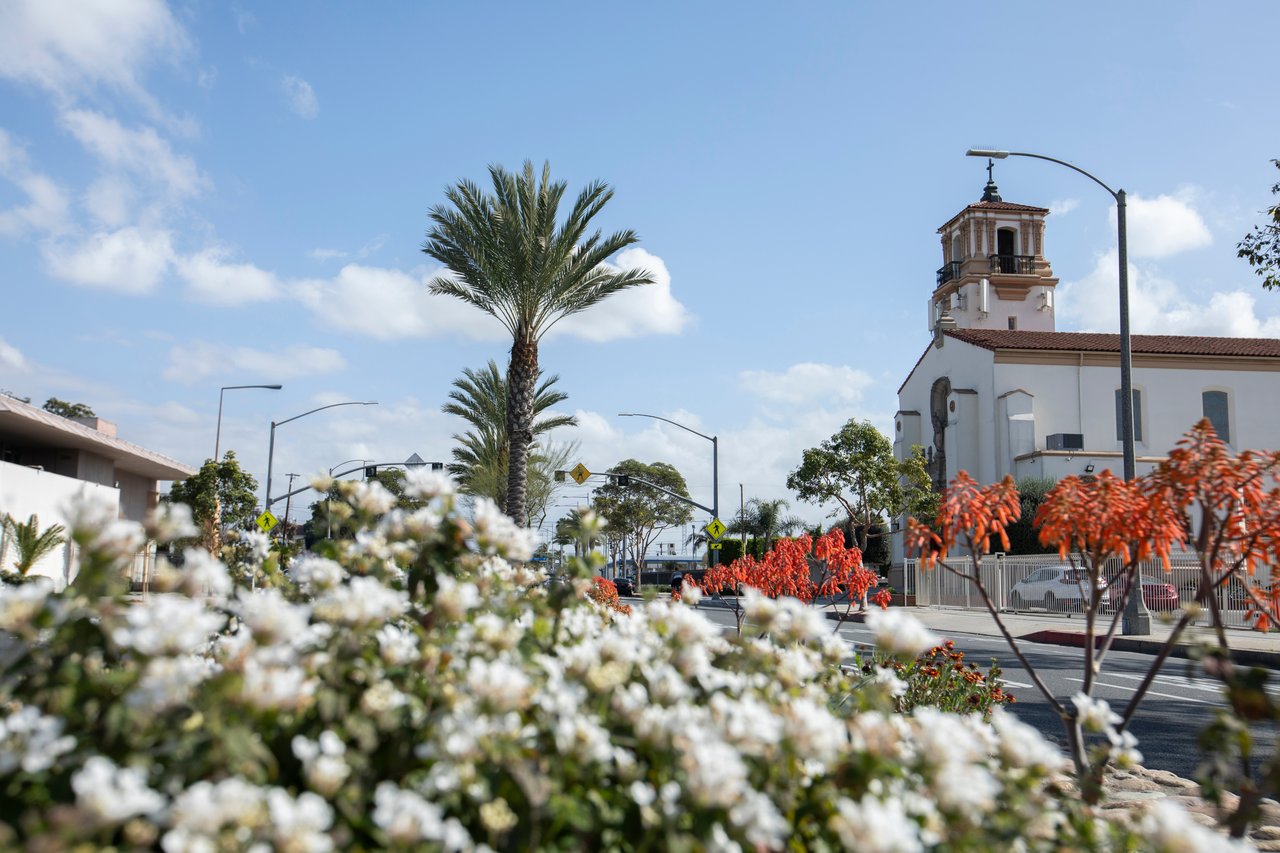 South Gate street with a white church and bell tower, palm trees, and flowering shrubs.