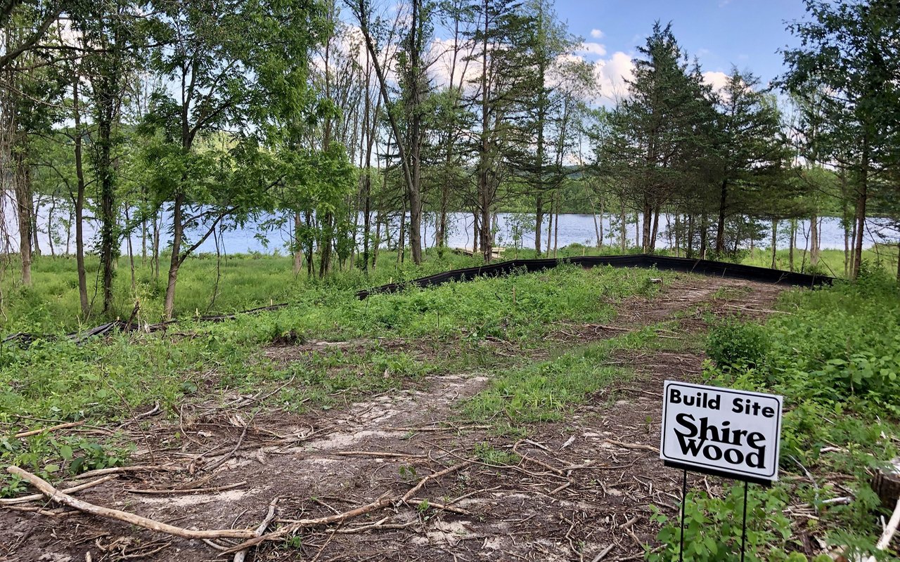 Wooded Shire Wood build site beside a lake, marked by a sign.