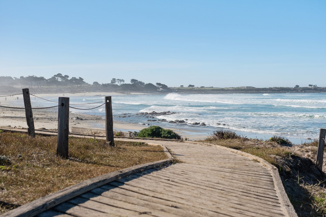 Wooden boardwalk with rope railing leading toward sandy Asilomar State Beach, with ocean waves and a coastal tree line in the background under a clear blue sky
