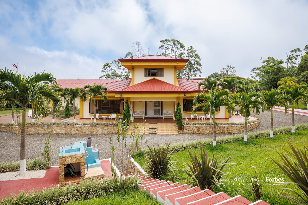Yellow house with red roofs, palm-lined grounds, and a small blue water feature.