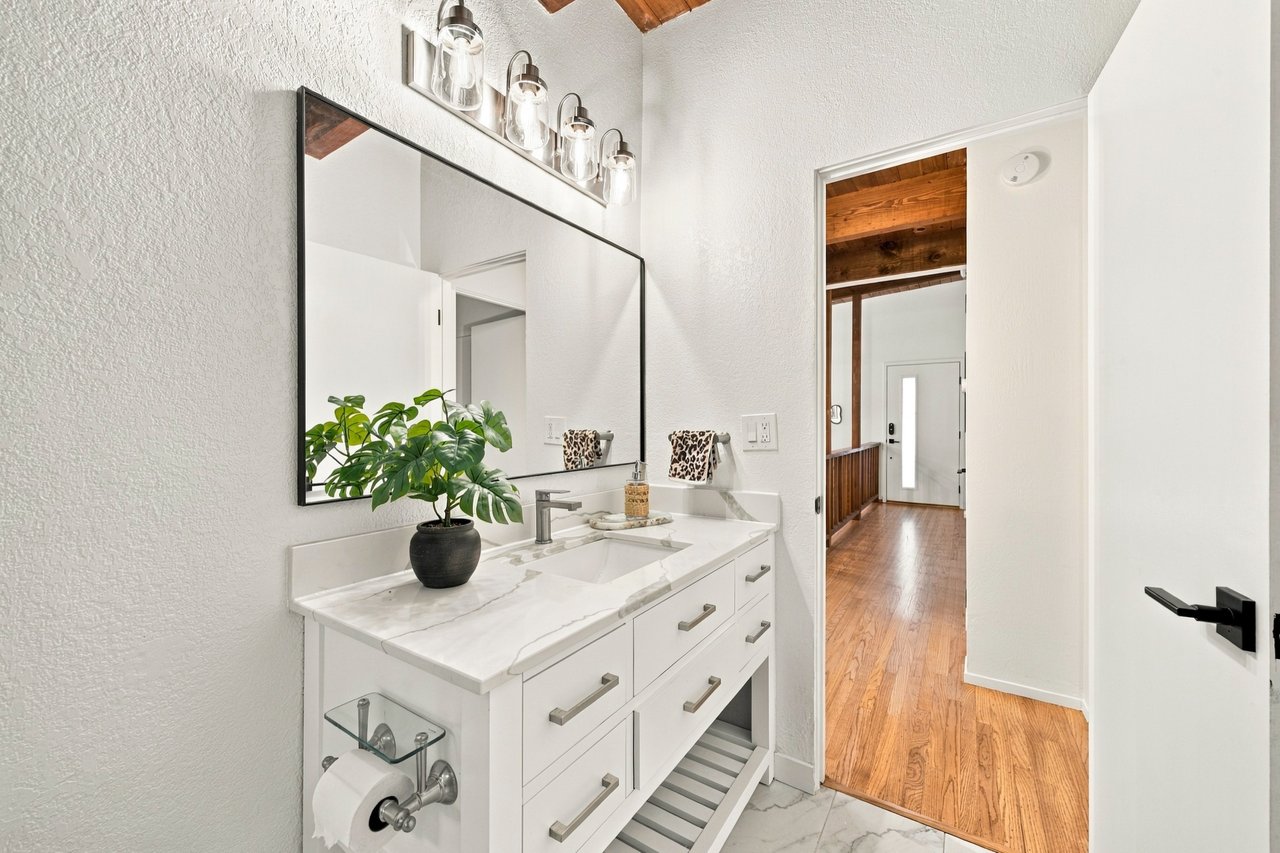 Bathroom vanity with a marble countertop, large mirror, and wood-floored hallway beyond.