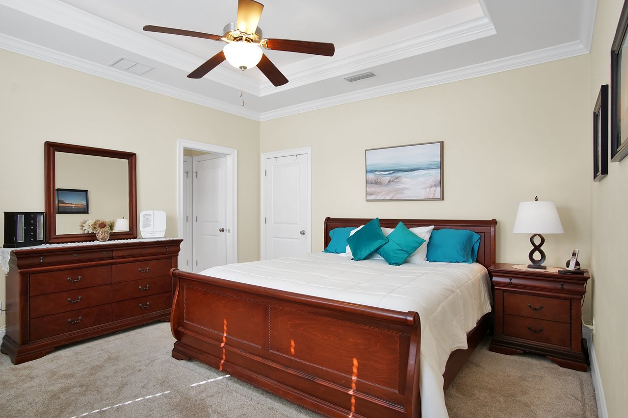 Spacious bedroom with a wooden bed and teal pillows. A ceiling fan is above, and a dresser with a mirror is on the left. The room feels cozy and elegant.