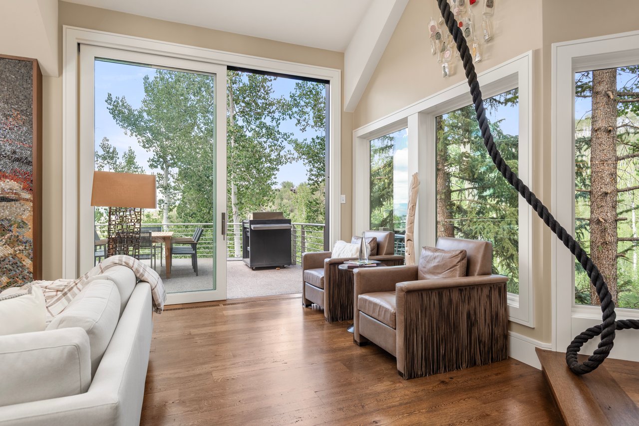 Living room with leather chairs beside large windows overlooking trees and a furnished deck.
