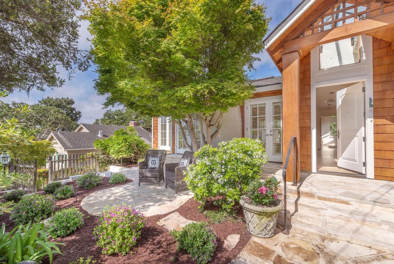 Home entrance with an open white door, stone patio, garden beds, and shaded seating.