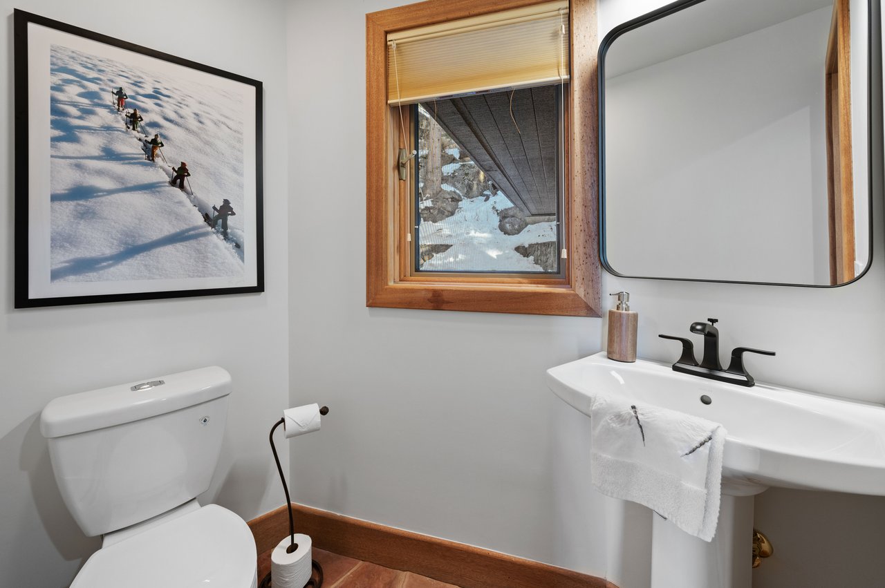 Bathroom with a pedestal sink, wood-trimmed window, and framed photograph of skiers.