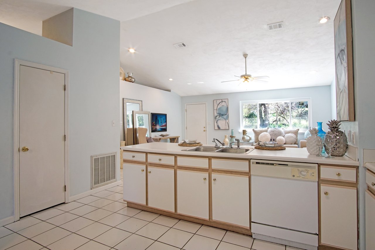 A bright, airy kitchen with white cabinets and countertops features a sink and dishwasher. Large windows with garden views create a serene atmosphere.