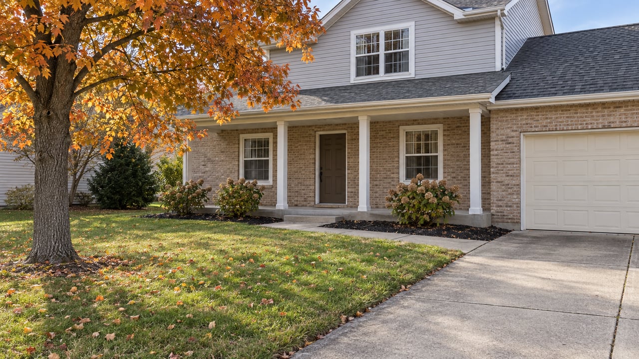 A modest brick-and-gray-sided two-story home with a concrete driveway, broad lawn and an amber-leaved sugar maple.