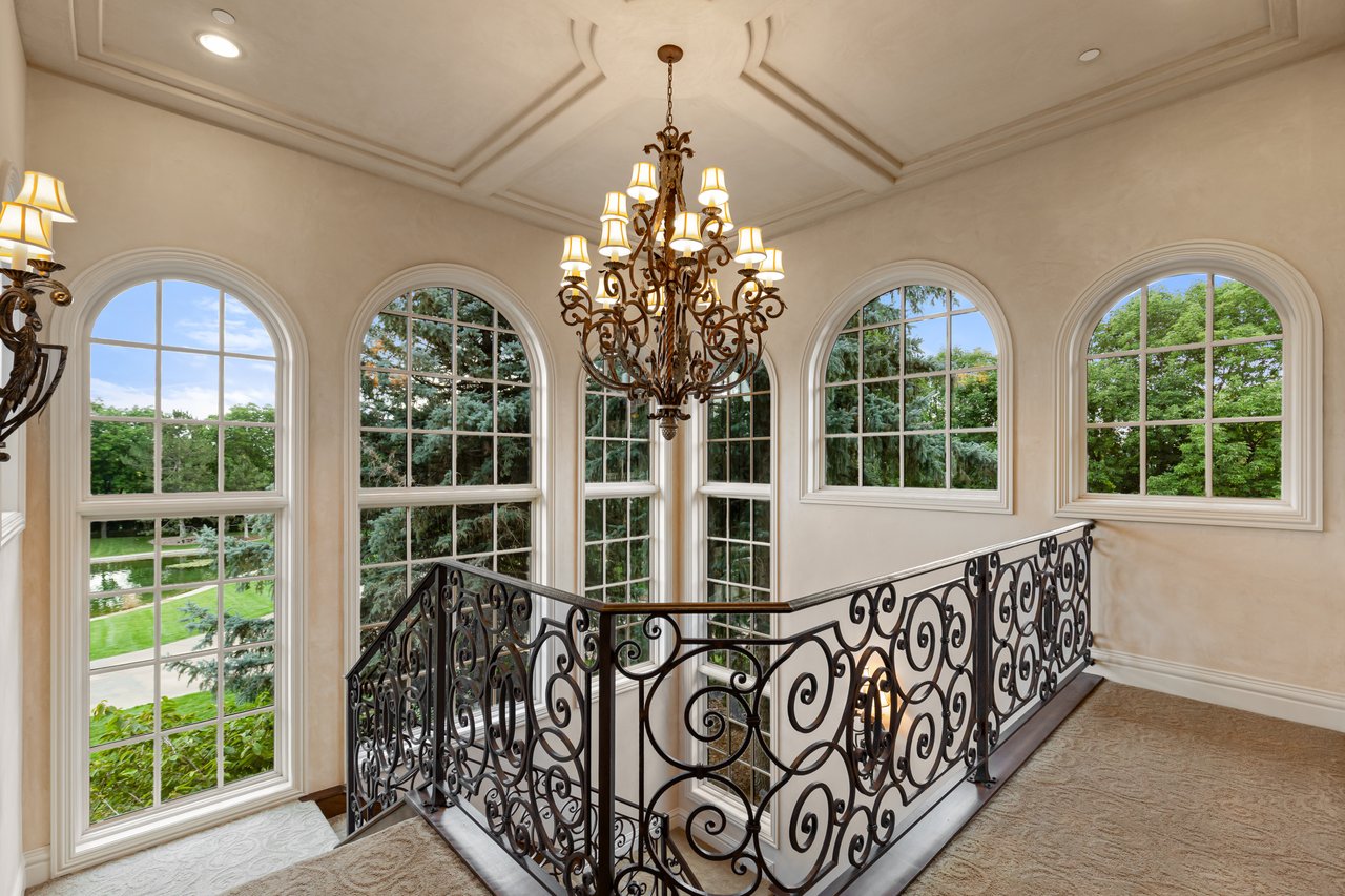 Upstairs staircase landing with ornate iron railing, chandelier, and tall arched windows overlooking trees.