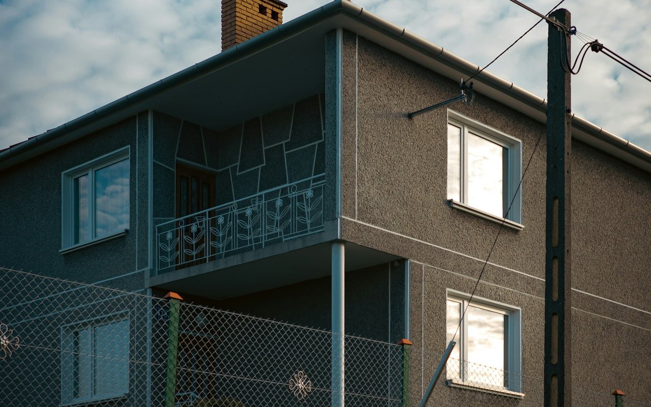 Two-story gray stucco home with a decorative balcony railing and chain-link fence.
