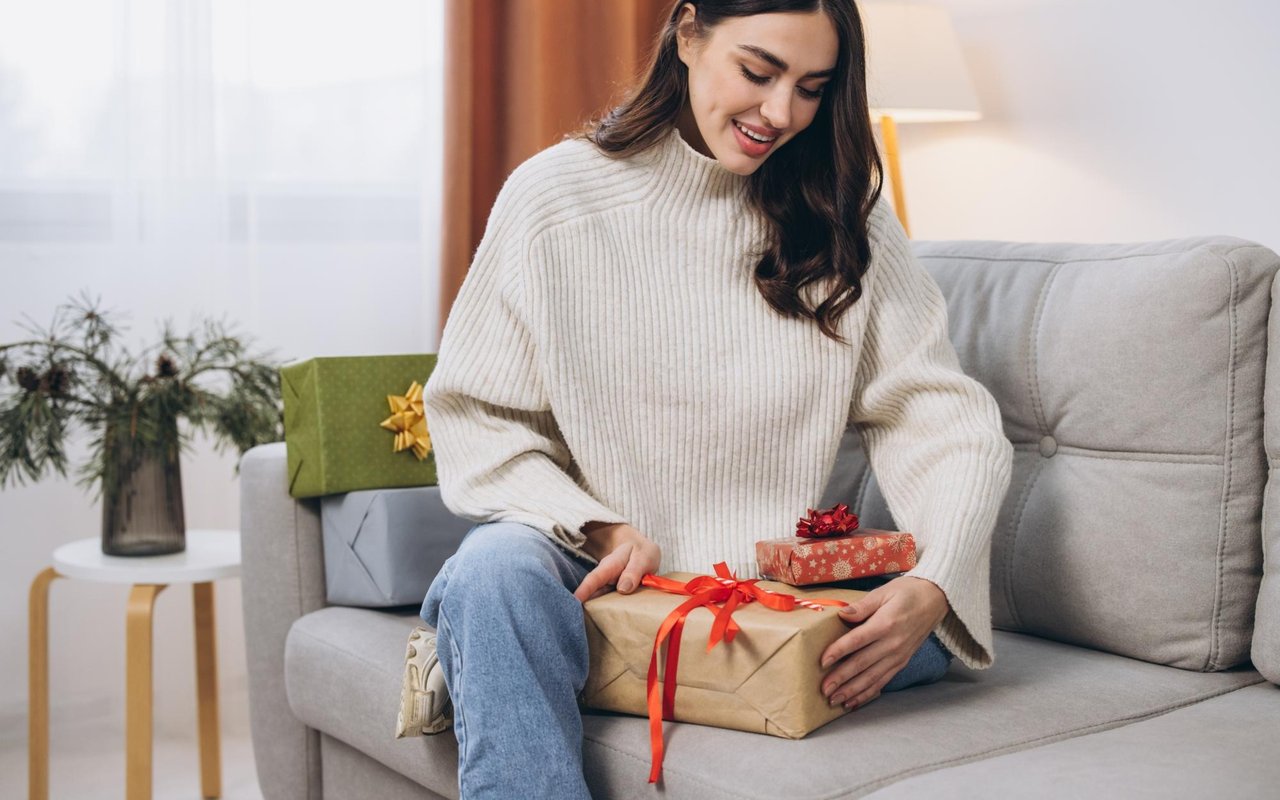 Woman opens a red-ribboned gift while sitting on a sofa beside other presents.