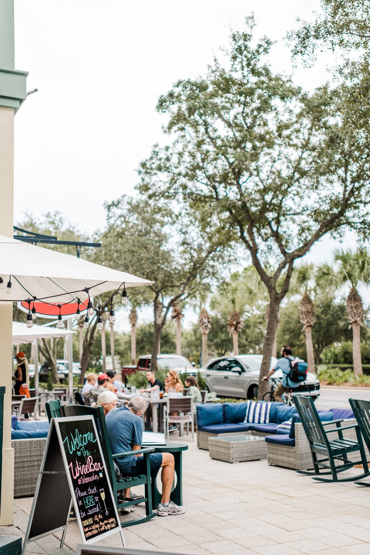 Live-oak-shaded town-center patio with a 'Wine Bar at WaterColor' chalkboard, market umbrellas and rocking chairs on 30A, Florida