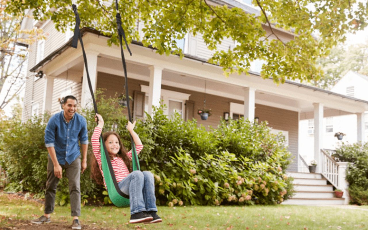 Two-story house with a covered porch and front-yard swing where a man watches a child.