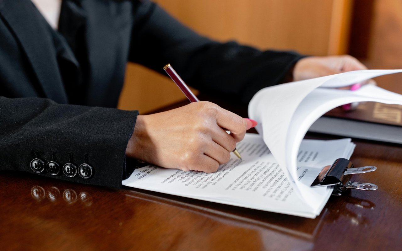 A person in a dark suit signs paperwork beside a clipboard and extra documents.