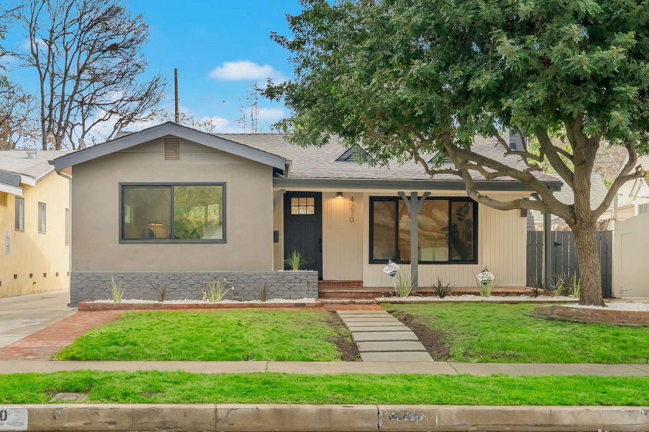 Single-story home with gray and cream siding, a curved walkway, green lawn, and large shade tree.