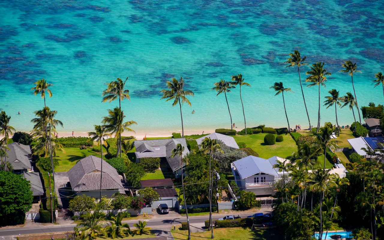 Coastal homes beside a sandy beach and turquoise water, framed by tall palm trees.