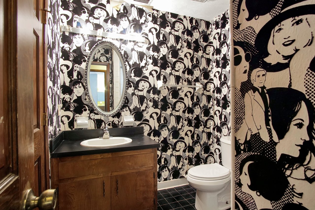 Bathroom with chic black-and-white pop art wallpaper featuring various faces. Oval mirror, wooden vanity, and white toilet create a retro vibe.