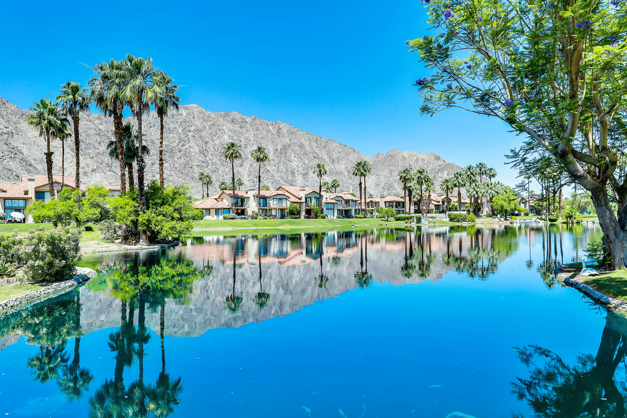Homes and palm trees reflected in a lake, with rugged mountains behind them.