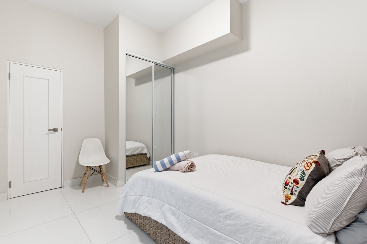 Furnished bedroom with a white bed, patterned pillows, mirrored closet doors, and a white chair.