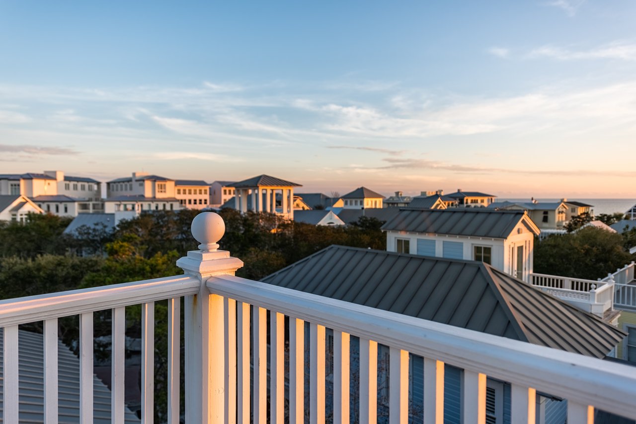 Balcony overlooking coastal homes and the ocean at sunset, with a white railing and metal roofs.