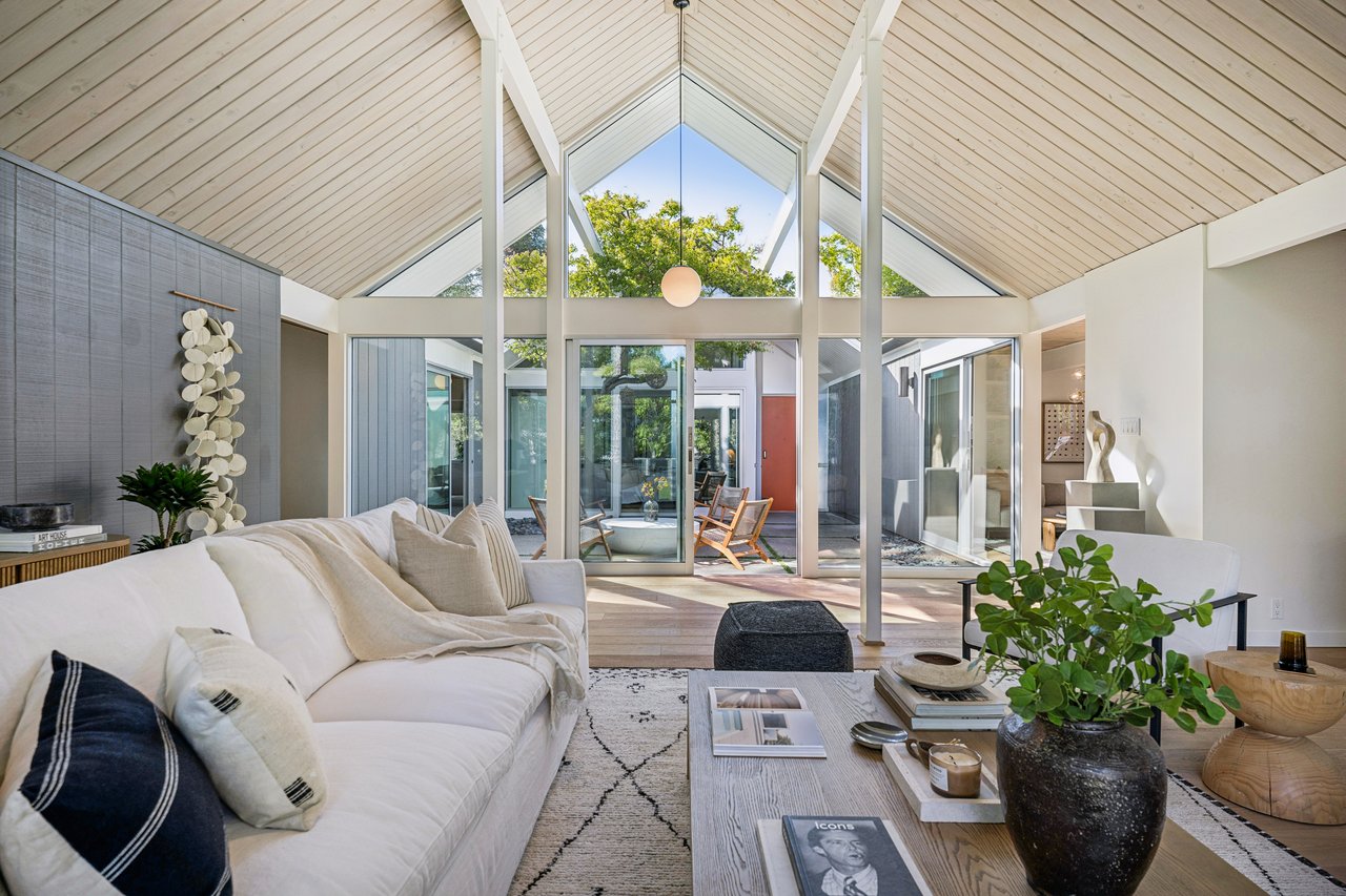 Vaulted living room with a white sofa, pale wood ceiling, and glass doors opening to a leafy courtyard.