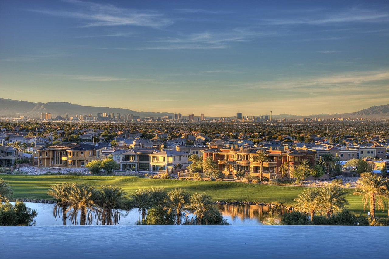 Upscale homes and golf course overlook the Las Vegas skyline, with mountains beyond and a pool in the foreground.