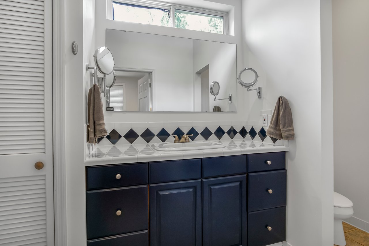 A white bathroom features a large mirror above a sink, with dark cabinets below. Two brown towels hang on either side, and diamond-pattern tiles add accents.