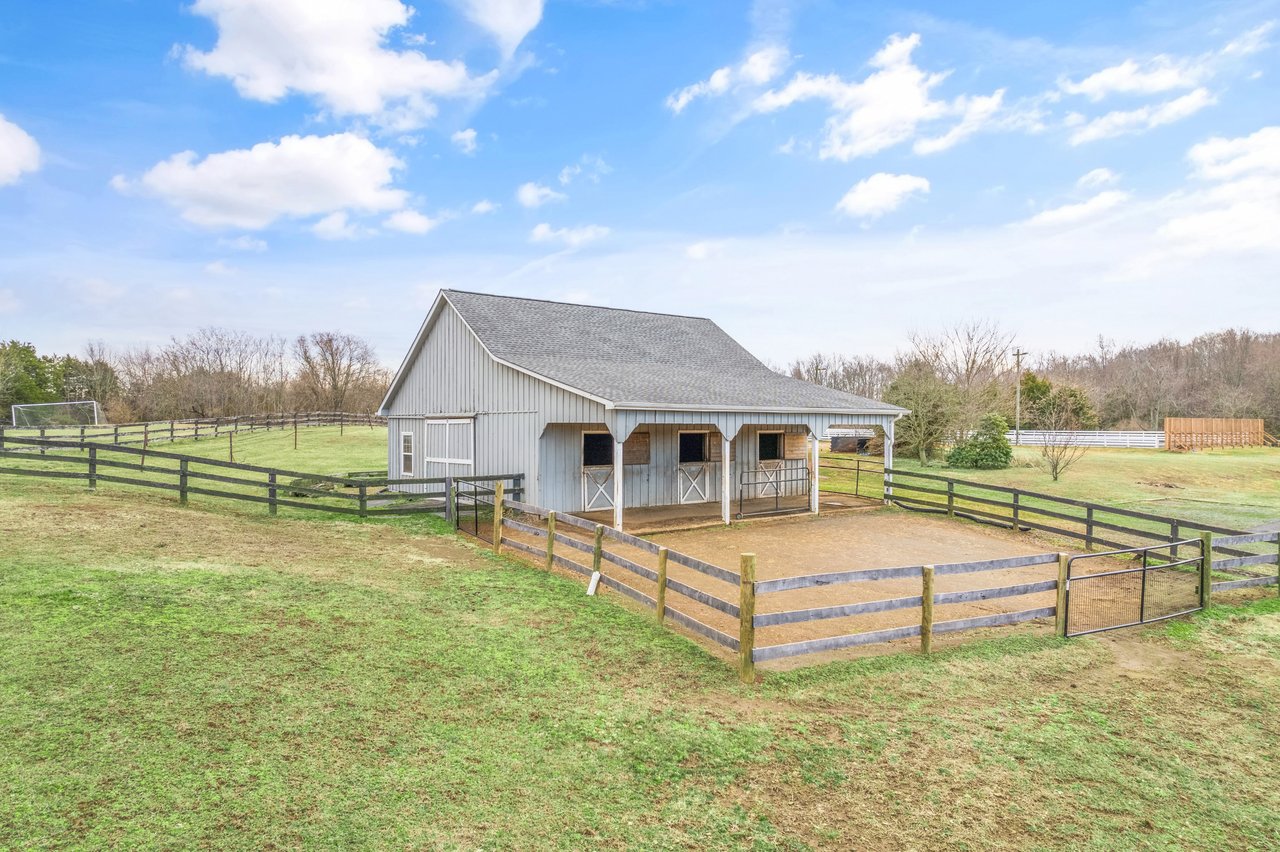 Light-gray horse barn with multiple stalls, fenced paddocks, and a dirt exercise area.