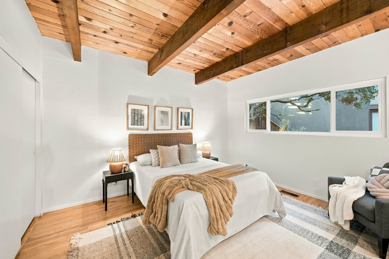 Bedroom with exposed wood-beam ceiling, a neatly made bed, and a long horizontal window.