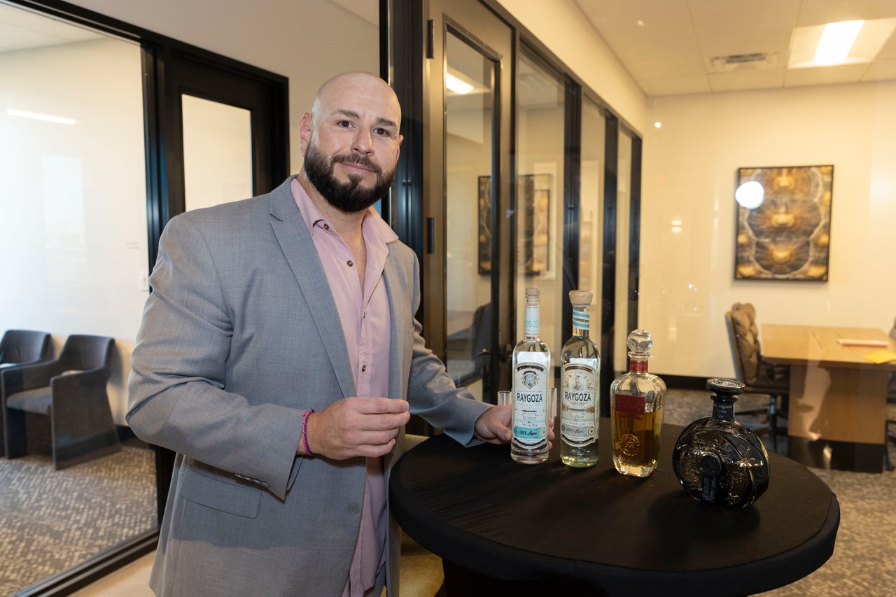 Bearded man in a gray suit presenting bottles of spirits at an indoor open house.
