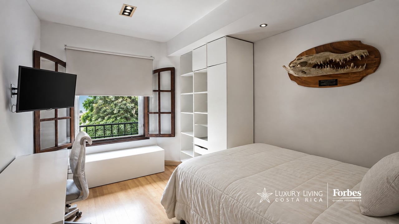 Bedroom with a white bed, built-in shelving, and a window seat beside a shaded window.