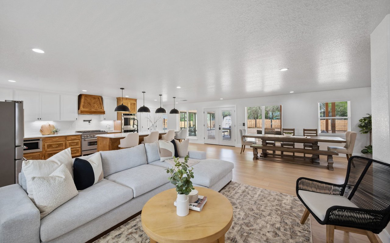 Open-plan kitchen and living room with a gray sectional, round coffee table, and long dining table by glass doors.