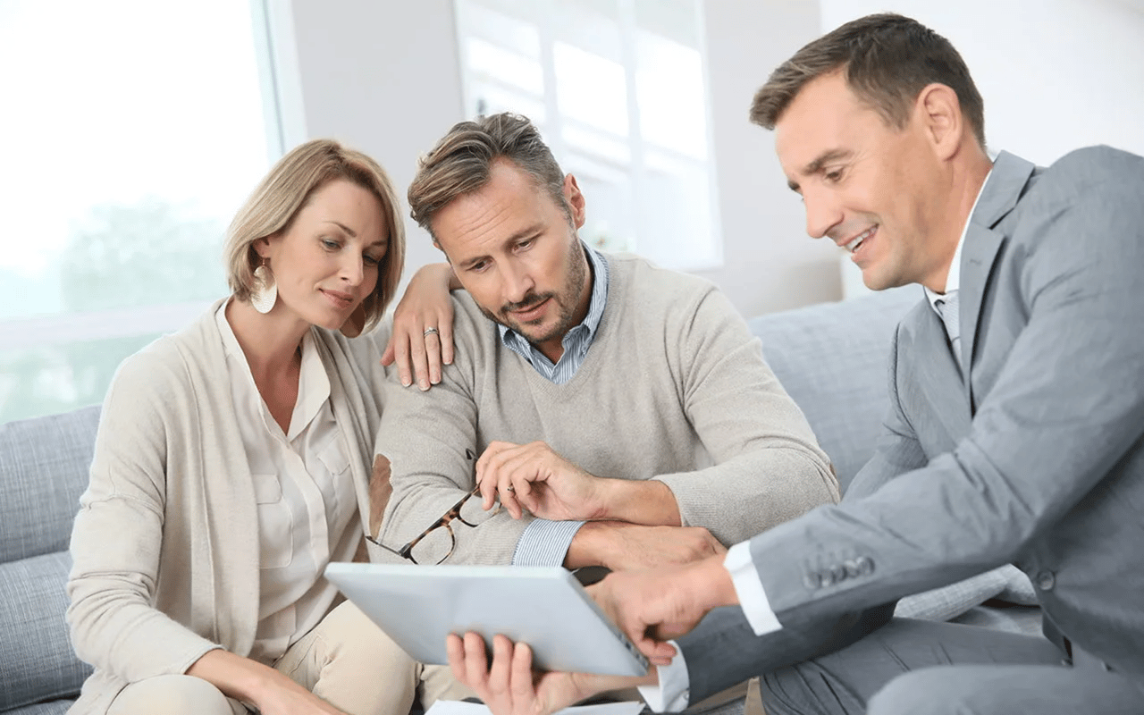 A real estate agent reviews a tablet with a couple seated on a sofa.