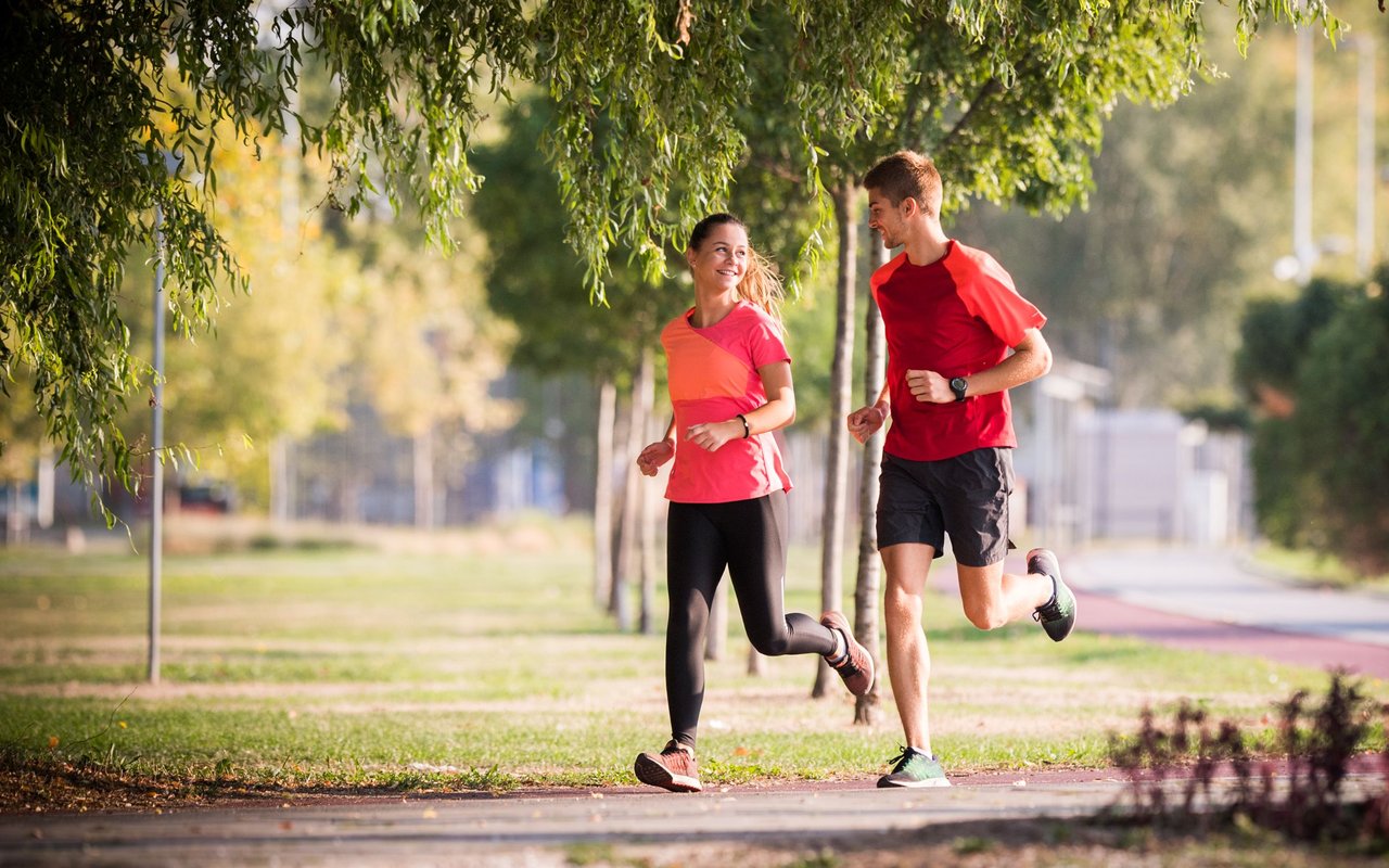 A man and woman jog together on a shaded path beneath leafy trees.