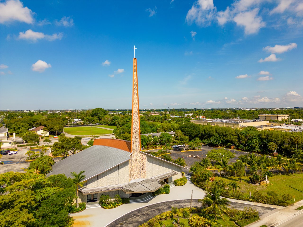 Aerial view of a church with a tall patterned spire, surrounded by trees and low-rise buildings.