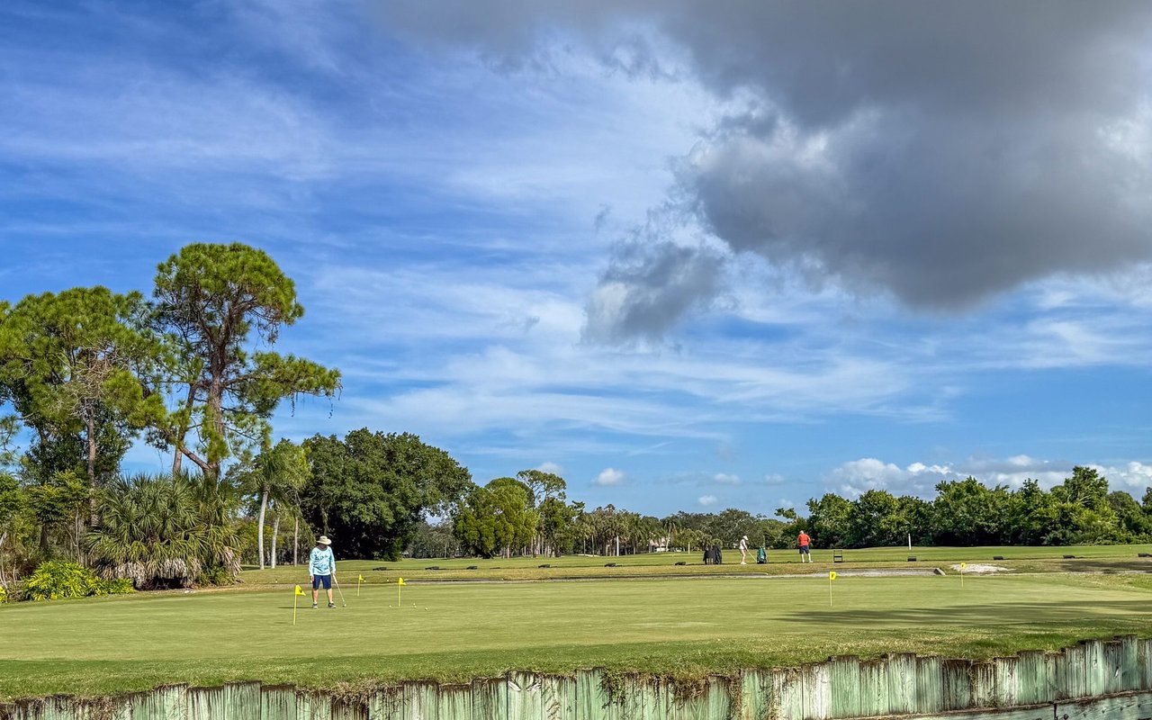 Golf course putting green with golfers and yellow flagsticks beneath a partly cloudy sky.