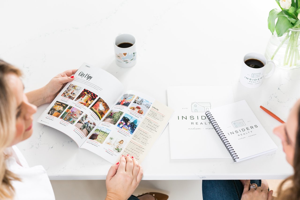 Cait Berry of Insiders Realty meeting with a relocating buyer at a clean white table, reviewing a Madison lifestyle magazine featuring local Instagram accounts, restaurants, and activities, while Insiders Realty welcome materials and a branded coffee mug rest on the table beside them.