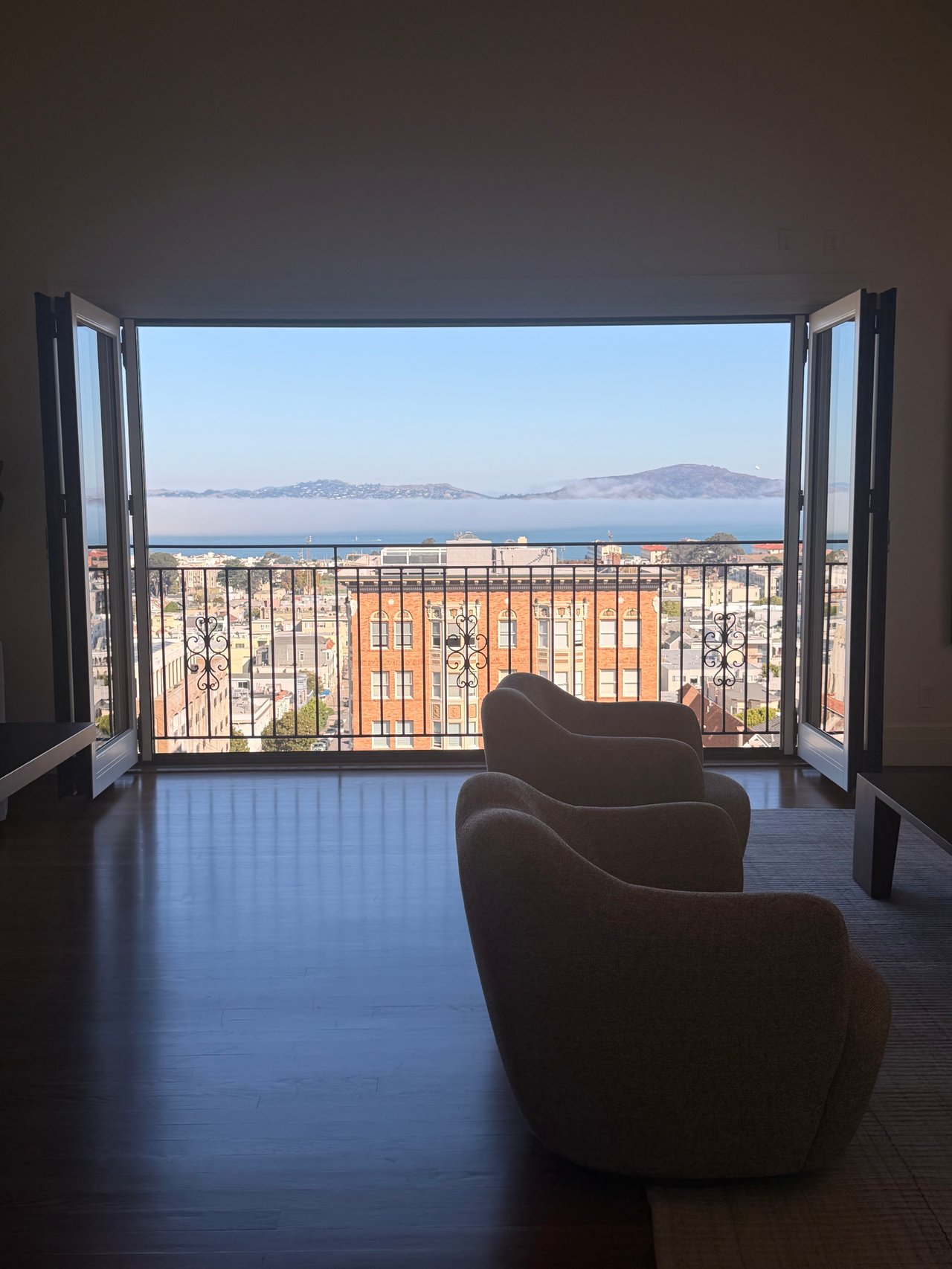 Living room with open windows overlooking San Francisco, the bay, and fog-covered hills.