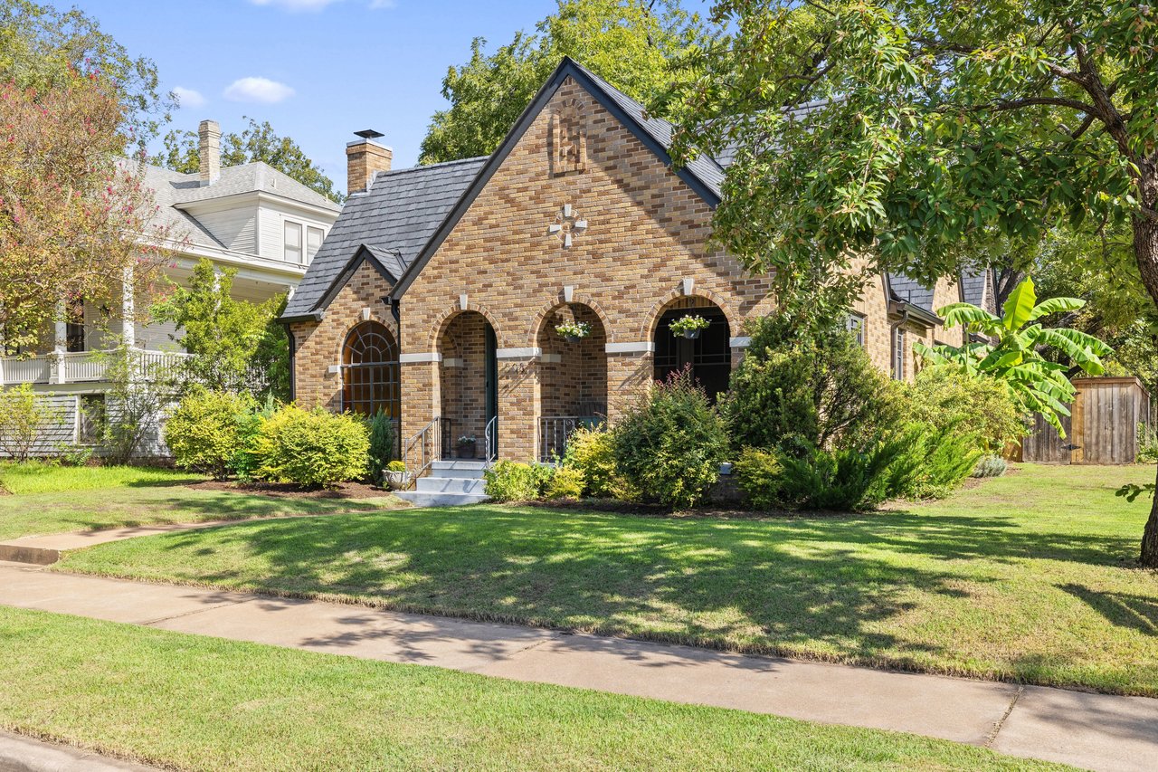 Brick house with a steep roof and arched porch, framed by mature trees and a green lawn.