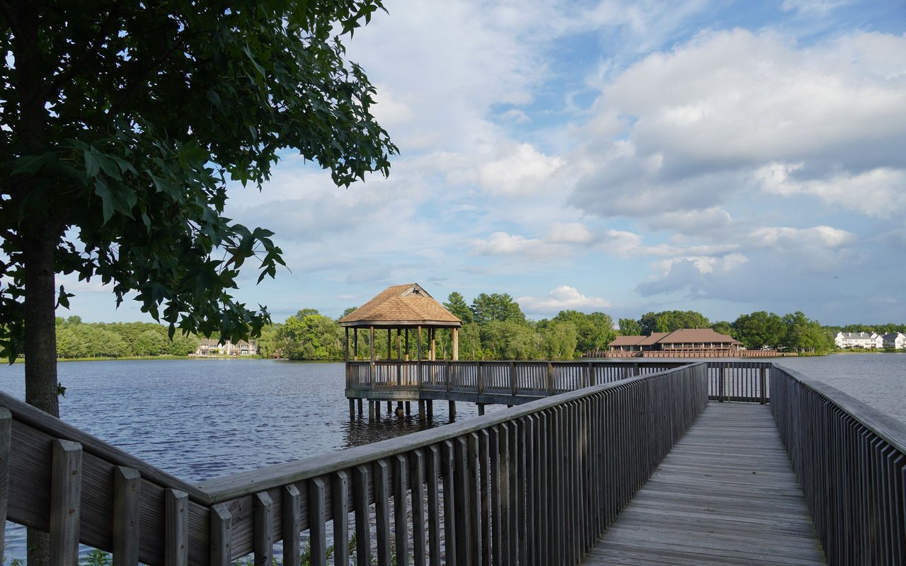 Wooden boardwalk crossing a lake toward a covered pavilion, with a tree-lined shore beyond.